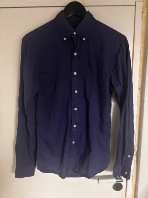 Mörkblå skjorta från Ralph Lauren - Klassisk mörkblå skjorta från Ralph Lauren med button-down krage och vita knappar. Skjortan har lång ärm och en liten röd broderad logga på bröstet. Tillverkad i mjuk bomull som känns skön mot huden. Perfekt för en stilren look.