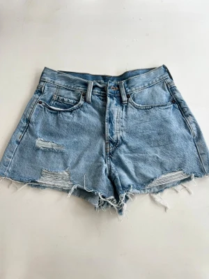 Blå denimshorts med fransig kant - Säljer ett par ljusblå denimshorts rå, fransig kant. Shortsen har slitna detaljer framtill, klassiska fem fickor och stängs med knapp och dragkedja. Perfekta för sommaren och ger en avslappnad vibe.