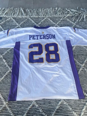NFL tröja Vikings Peterson #28 - Säljer en vit och lila NFL-tröja från Reebok med nummer 28 och namnet Peterson på ryggen. Tröjan har gula detaljer och korta ärmar. Perfekt för dig som gillar amerikansk fotboll/minnesota Vikings och vill sticka ut på matchen. Mycket bra skick, loggan på insidan av nacken har försvunnit lite grann men påverkar inte