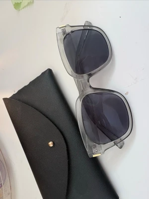 solglasögon - Snygga solglasögon med transparent, fyrkantig båge och mörka glas. Glasögonen har en modern oversized look och små gulddetaljer vid gångjärnen. Perfekta för dig som vill sticka ut och addera en cool vibe till din outfit. Svart fodral medföljer.