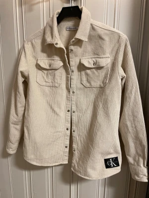 Beige manchester skjorta/ overshirt från Calvin Klein - Säljer en beige overshirt i manchester från Calvin Klein. Skjortan har långa ärmar, två bröstfickor med lock och knappar framtill. CK-logga nertill på ena sidan ger en clean och trendig look. Stl 14 / XS