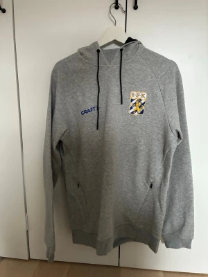 Grå IFK Göteborg hoodie från Craft (OANVÄND) - Snygg grå hoodie från Craft med IFK Göteborgs klubbmärke broderat på bröstet. Hoodien har svarta dragsnören, känguruficka och ribbade muddar. Tillverkad i mjukt sweatshirtmaterial, perfekt för dig som vill representera laget. Tröjan kostar 1000 kr ny så 200 kr är en bra deal för köparen av denna oanvända tröja!