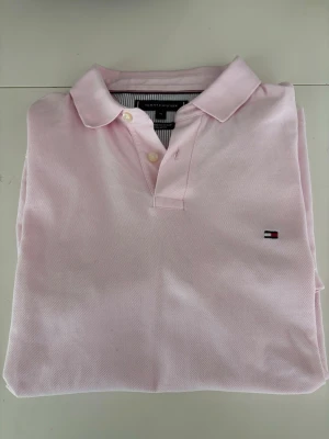 Ljusrosa pikétröja från Tommy Hilfiger - Säljer en ljusrosa pikétröja från Tommy Hilfiger med klassisk krage och två knappar framtill. Tröjan har Hilfiger-logotypen broderad på bröstet och är gjord i mjuk bomull.