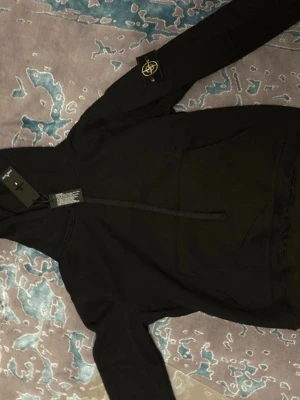 Svart hoodie från Stone Island - Storlek M eller mindre L. Säljer en svart hoodie från Stone Island med klassisk patch på vänster ärm och stor magficka framtill. Perfekt för dig som gillar streetwear och vill ha en stilren look.