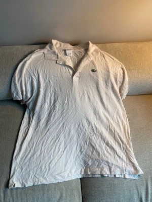 Vit pikétröja från Lacoste - Klassisk vit pikétröja från Lacoste med den ikoniska gröna krokodilloggan på bröstet. Tröjan har korta ärmar, krage och knäppning framtill. Perfekt till en avslappnad och stilren look.