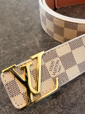Louis Vuitton rutigt bälte med LV-spänne - Snyggt bälte från Louis Vuitton med klassiskt rutigt mönster i vitt och beige. Bältet har ett stort guldfärgat LV-spänne framtill och är gjort i skinn. Perfekt accessoar för att lyfta din outfit.