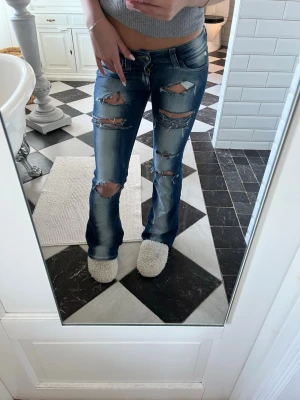 Blå slitna bootcut jeans - Säljer ett par blå bootcut jeans med flera stora slitningar och hål på både lår och knän. Jeansen har en klassisk femficksmodell och är tillverkade i denim med tvättad effekt för en cool, avslappnad look. Jag är 170 för referens 