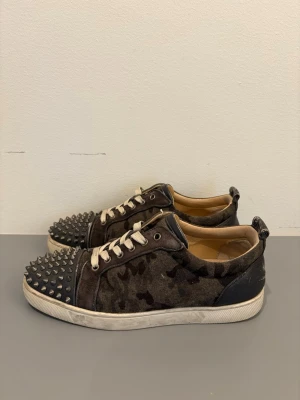 Christian Louboutin sneakers - Unika sneakers från Christian Louboutin i mörkgrönt camouflagemönster med svarta läderdetaljer och silverfärgade nitar på tån. Skorna har vita snören och en vit sula. Perfekta för dig som vill sticka ut med din stil.