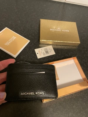 Svart korthållare Michael Kors - Stilren svart korthållare från Michael Kors i präglat läder. Har flera fack för kort och silver logga framtill. Perfekt för dig som vill ha ett smidigt och snyggt sätt att hålla koll på dina kort.