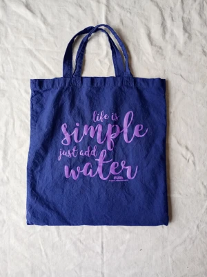 Blå shoppingväska med text - Snygg blå shoppingväska i tyg med trycket 'life is simple just add water' i lila text på framsidan. Väskan har två långa handtag och är perfekt för att bära med sig böcker, mat eller annat smått och gott. Enkel och stilren design.
