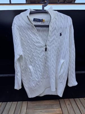 Vit kabelstickad tröja Polo Ralph Lauren🙌🤩 - Vit kabelstickad tröja från Polo Ralph Lauren med halv dragkedja och hög krage. Tröjan har två fickor framtill och den klassiska mörkblå logotypen broderad på bröstet. Ribbstickade muddar vid ärmslut och nederkant. Perfekt för en stilren look.🙌🤩