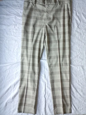 Beige rutiga kostymbyxor - Snygga beige kostymbyxor med rutigt mönster och raka ben. Byxorna har bälteshällor och sidofickor, perfekta för en clean och stilren look. Passar dig som gillar klassisk men trendig stil.