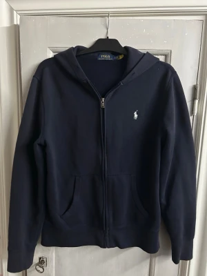 Polo Ralph Lauren zip hoodie - Hej! Säljer en blå Polo Ralph Lauren hoodie med dragkedja i storlek M.                         Säljer den pågrund av att jag växt ur den, men hoodien är i ett fint skick!  !OBS! Tröjan är äkta och har ett äkthets bevis i nacken om man scannar QR koden.  Skriv gärna vid frågor! Axelbredd ca 45cm längd ca 67cm
