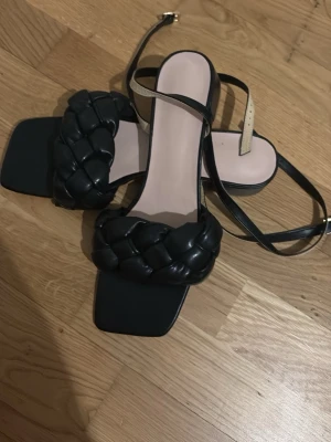 Svarta flätade sandaler med rem - Stilrena svarta sandaler med bred, flätad rem över foten och smal ankelrem med spänne. Sandalerna har fyrkantig tå och slät sula. Tillverkade i konstläder med beige innersula för en modern look.