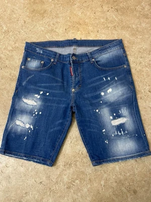 Blå jeansshorts med slitningar - Säljer ett par blå jeansshorts med slitna detaljer och fräsch tvätt. Klassisk femficksmodell med knappgylf och bälteshällor. Perfekta för en avslappnad stil i sommar.