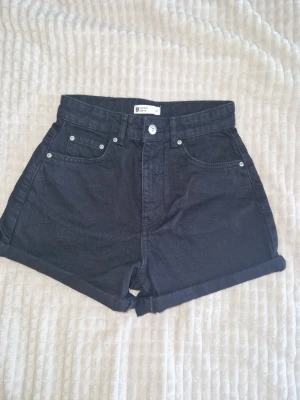 Svarta high waist jeansshorts Gina Tricot - Snygga svarta jeansshorts från Gina Tricot med hög midja och klassisk femficksmodell. Shortsen har uppvikta benslut och stängs med knapp och dragkedja. Perfekta till sommaren för en avslappnad och trendig look. Sitter som ett smäck😍🍑 