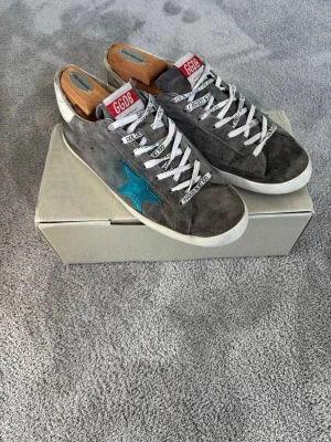 Grå Golden Goose sneakers med blå stjärna - Säljer ett par grå sneakers från Golden Goose med vit sula, vita skosnören och en blå stjärna på sidan. Skorna har GGDB-logga på plösen och text på snörena. Ovandelen är i mocka och har en avslappnad, vintage look. Kommer det ett par vita skosnören och ett par svarta.