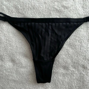 Svarta stringtrosor med ränder - Säljer ett par svarta stringtrosor med tunna, diskreta ränder i tyget. Trosorna har dubbla tunna band i midjan och är tillverkade i ett lätt och stretchigt material som ger en bekväm passform. Perfekta för dig som gillar en stilren och enkel look. Ny pris 180kr