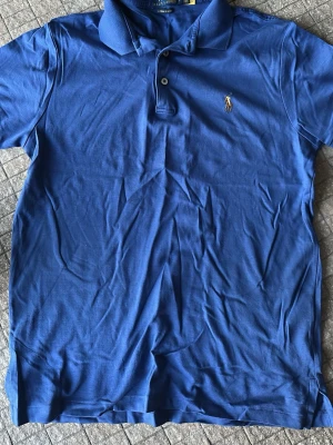 Blå piké från Polo Ralph Lauren - Klassisk blå pikétröja från Polo Ralph Lauren med broderad logga på bröstet. Tröjan har korta ärmar, krage och knappar framtill. Tillverkad i mjuk supima cotton och har en slim passform som sitter snyggt. Använt 2-3 ggr. Toppen skick.