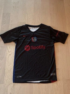 Fc barcelona tröja - Snygg svart FC Barcelona matchtröja från Nike med klubbmärke och röd Nike-logga på bröstet. Spotify-logga tryckt framtill. Tröjan har V-ringning och korta ärmar, tillverkad i ett lätt och ventilerande funktionsmaterial. Ny skick.