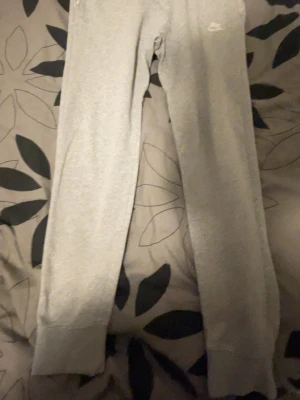 Gråa mjukisbyxor från Nike - Säljer ett par klassiska gråa mjukisbyxor från Nike med dragsko i midjan och ribbade muddar vid bensluten. Byxorna har en enkel och clean look med Nike-logga broderad på benet. Perfekta för chill dagar eller när du vill vara bekväm.