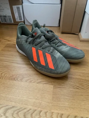 Adidas grå fotbollsskor med orange detaljer - Snygga fotbollsskor från Adidas i grått med de klassiska tre ränderna i orange på sidan. Skorna har snörning, låg profil och ovandel i syntetmaterial med textur för bättre bollkontroll. Perfekta för inomhusspel och snabba vändningar på planen.