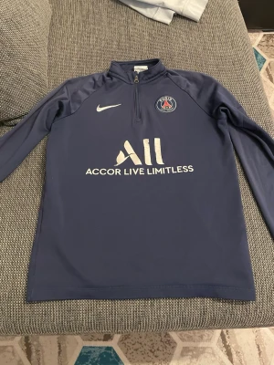 Blå PSG träningströja från Nike - Mörkblå långärmad träningströja från Nike med PSG-logga och vit sponsorprint på bröstet. Tröjan har halv dragkedja framtill och är tillverkad i ett stretchigt funktionsmaterial som passar perfekt till fotbollsträning.
