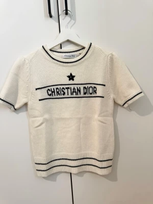 Vit stickad tröja  - Vit stickad tröja  med svarta detaljer och logga framtill. Tröjan har korta ärmar, rund halsringning och svarta ränder vid ärmslut, hals och nederkant. Tillverkad i Italien och har en klassisk, stilren design.