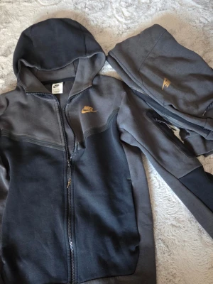 Nike grå och svart loungewear-set - Nike loungewear-set med zip-hoodie och matchande byxor i mörkgrå och svart färg. Setet har guldfärgade Nike-loggor på både tröja och byxa. Tillverkat i mjukt bomullsmaterial, perfekt för chill dagar hemma. Hoodien har dragkedja och fickor.