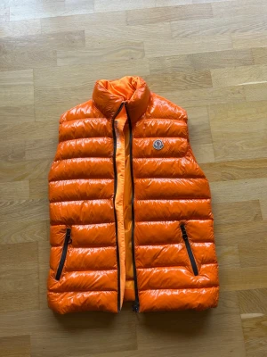 Orange dunväst från Moncler - Snygg orange dunväst från Moncler med glansig finish och ståkrage. Västen har två svarta dragkedjefickor framtill och Moncler-logga på bröstet. Perfekt för lager-på-lager och ger en cool streetwear-look. Går inte att stänga dragkedjan men går lätt att fixa!
