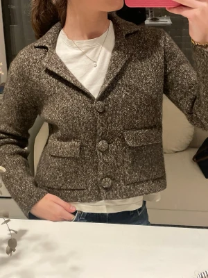 Brun stickad kofta med knappar - En snygg brunmelerad stickad kofta med krage och stora knappar framtill. Koftan har två fickor på framsidan och är kort i modellen. Perfekt att slänga över en t-shirt för en cool och avslappnad look.