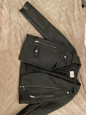 Svart skinnjacka från Zara - Snygg svart skinnjacka från Zara med klassisk bikerstil. Jackan har silvriga dragkedjor, snedställd frontzip och fickor med tryckknapp. Perfekt för dig som vill ha en stilren och trendig look.