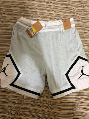 Jordan shorts - Snygga vita Jordan shorts med svarta detaljer och klassisk Jumpman-logga på benen. Shortsen har elastisk midja med snörning och är tillverkade i ett ventilerande meshmaterial, perfekt för sport eller chill. Stilsäker design med sportig känsla.