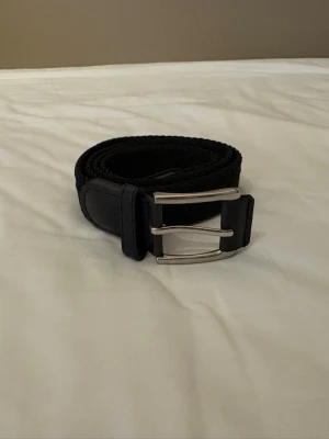 Svart flätat bälte med silverfärgat spänne - Snyggt svart flätat bälte med ett stilrent silverfärgat spänne. Bältet har detaljer i skinn och passar perfekt till jeans eller byxor för en clean look. Enkel att justera tack vare det flexibla materialet och den klassiska designen.