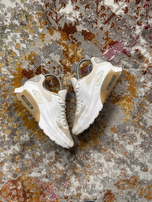 Nike Air Max 90 vita/beige sneakers - Snygga Nike Air Max 90 sneakers i vitt och beige med klassisk synlig Air-sula. Ovandelen är i mesh och läder med detaljer i mocka. Skorna har en chunky sula och ikoniska Air Max-loggor på sidan och hälen. Perfekta för dig som gillar stilrena och sportiga sneakers.