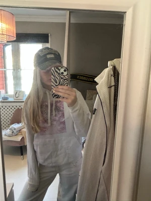 The cool elephant hoodie - Säljer en vit hoodie med huva och dragsnören. På bröstet finns ett stort konstnärligt tryck i rosa och lila toner som föreställer ett ansikte och blommor. Hoodien har en avslappnad passform och är tillverkad i mjukt bomullsmaterial. Perfekt för en chill och kreativ stil.