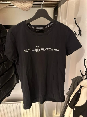 Svart Sail Racing t-shirt - Snygg svart t-shirt från Sail Racing med vit logga tryckt på bröstet och en mindre logga i nacken. Klassisk rund hals och korta ärmar. Perfekt till jeans eller shorts för en clean och sportig look.