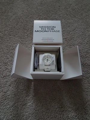 Omega x Swatch MoonSwatch Moonphase - Snygg vit Omega x Swatch MoonSwatch Mission to the Moonphase med biokeramiskt material. Klockan har kronograf, tydlig urtavla och vit rem med Omega-logga. Perfekt för dig som gillar stilrena accessoarer och vill sticka ut med en unik design.