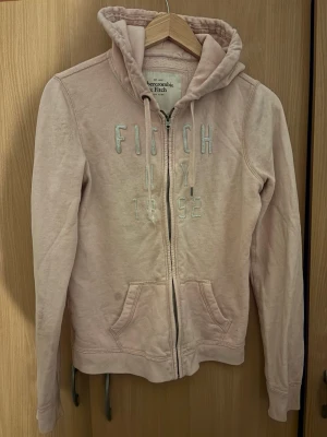 Ljusrosa hoodie Abercrombie & Fitch - Ljusrosa hoodie från Abercrombie & Fitch med dragkedja, broderad text 'FITCH NY 1892' på bröstet och två fickor framtill. Mjuk bomullsmix och klassisk huva med snören. Perfekt för en avslappnad och trendig look.