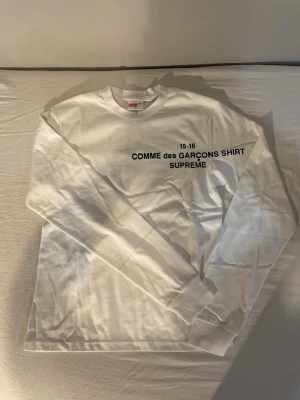 Vit långärmad Supreme x Comme des Garçons - Vit långärmad topp från Supreme och Comme des Garçons med svart tryck på bröstet. Klassisk crewneck och ribbade muddar vid ärmslut. Perfekt för dig som gillar streetwear och stilrena plagg.