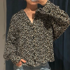 Leopardmönstrad skjorta med knappar - Säljer en snygg leopardmönstrad skjorta med v-ringning och knappar framtill. Skjortan har lång ärm och en loose passform, perfekt för dig som vill sticka ut lite extra. Materialet känns mjukt och lätt, troligen polyester. Passar till jeans eller kjol för en cool look.