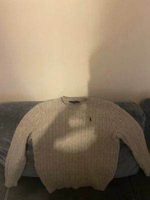 Beige kabelstickad tröja Polo Ralph Lauren - Kabelstickad beige tröja från Polo Ralph Lauren med rund halsringning och klassisk svart broderad logga på bröstet. Tröjan har långa ärmar och ribbade muddar. Perfekt för dig som gillar stilrena och tidlösa plagg. Pris kan diskuteras skicka pm!