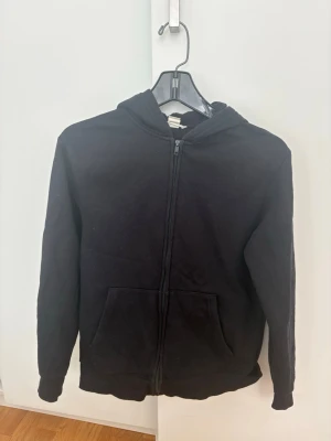 Svart zip hoodie med huva - En klassisk svart hoodie med dragkedja framtill och två fickor. Hoodien har en huva och ribbade muddar vid ärmar och nederkant. Perfekt för en avslappnad stil och enkel att matcha med det mesta. Materialet känns mjukt och bekvämt, troligen bomullsblandning.