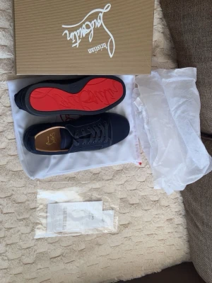 Svarta Louboutin sneakers med röd sula - Snygga svarta sneakers från Christian Louboutin med ikonisk röd sula och diskret logga på innersulan. Skorna har klassisk låg profil, svarta skosnören och ovandel i textil. Perfekta för dig som vill sticka ut med stilrena detaljer.pris kan diskuteras !!!!