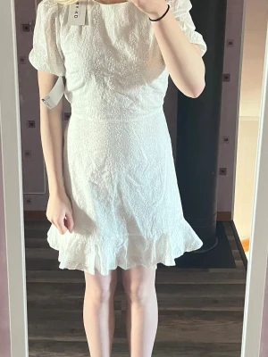 Vit kort klänning med volang - Söt studentklänning från NA-KD med broderade blommor, puffärmar och volangkant nedtill. Klänningen har rund halsringning och är tillverkad i bomull, vilket gör den både luftig och bekväm. Perfekt för sommarens alla tillfällen!