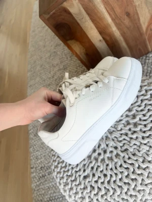 Vita sneakers från Gant - Stilrena vita sneakers från Gant med klassisk låg profil och vita snören. Skorna har en diskret logga på sidan och är tillverkade i slätt läder med en vit gummisula. Perfekta för en clean och fräsch look.