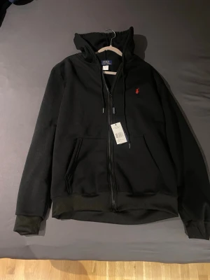 Svart zip up från Ralph Lauren - Säljer en svart hoodie från Ralph Lauren med dragkedja och huva. Klassisk design med två fickor framtill och den ikoniska röda logotypen broderad på bröstet. Tillverkad i mjukt bomullsmaterial och har ribbade muddar vid ärmslut och nederkant.