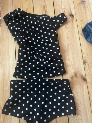 Svart prickigt pyjamas-set - Säljer ett svart set med vita prickar. Setet består av en kortärmad topp och matchande shorts. Materialet känns mjukt och bekvämt. Inget synligt märke på plagget.
