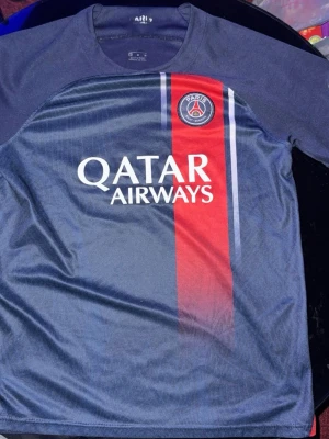 PSG matchtröja Mbappé nr 7 - Snygg mörkblå PSG matchtröja med röd och vit rand på framsidan, Qatar Airways-tryck och klubbmärke. På ryggen står det Mbappé och nummer 7. Perfekt för fotbollsfans! Tillverkad i lätt och ventilerande material.