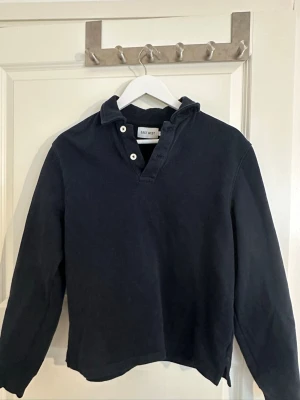 Mörkblå henley från East West - Snygg mörkblå henley från East West med klassisk krage och tre vita knappar vid halsen. Skjortan har lång ärm och är gjord i mjuk bomull, perfekt för en avslappnad och stilren look.